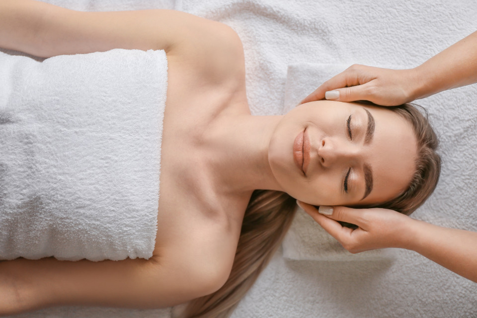 An esthetician’s hands resting on a client’s temples during a facial, the client lying on white towels with her eyes closed