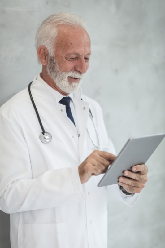 Dr. Alan Brekke, a gray-bearded doctor in a white coat, reading a tablet.