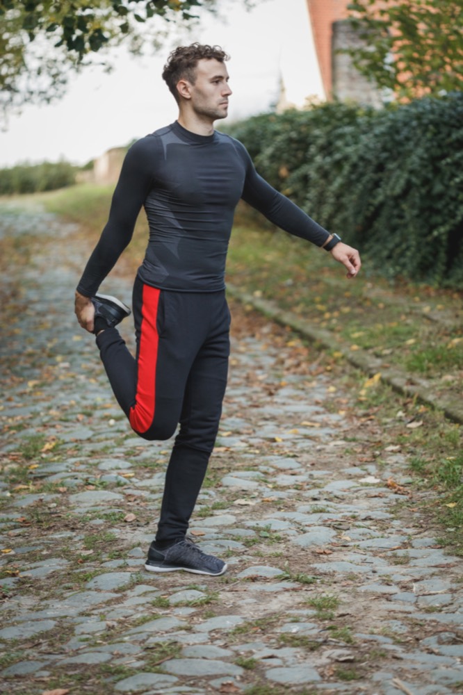 A runner holds a standing quad stretch on a park path.