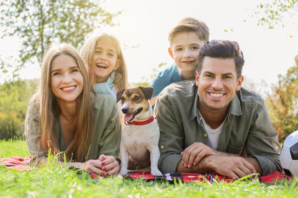 A mother, father, daughter and son lie on the grass with their dog, all smiling at the camera