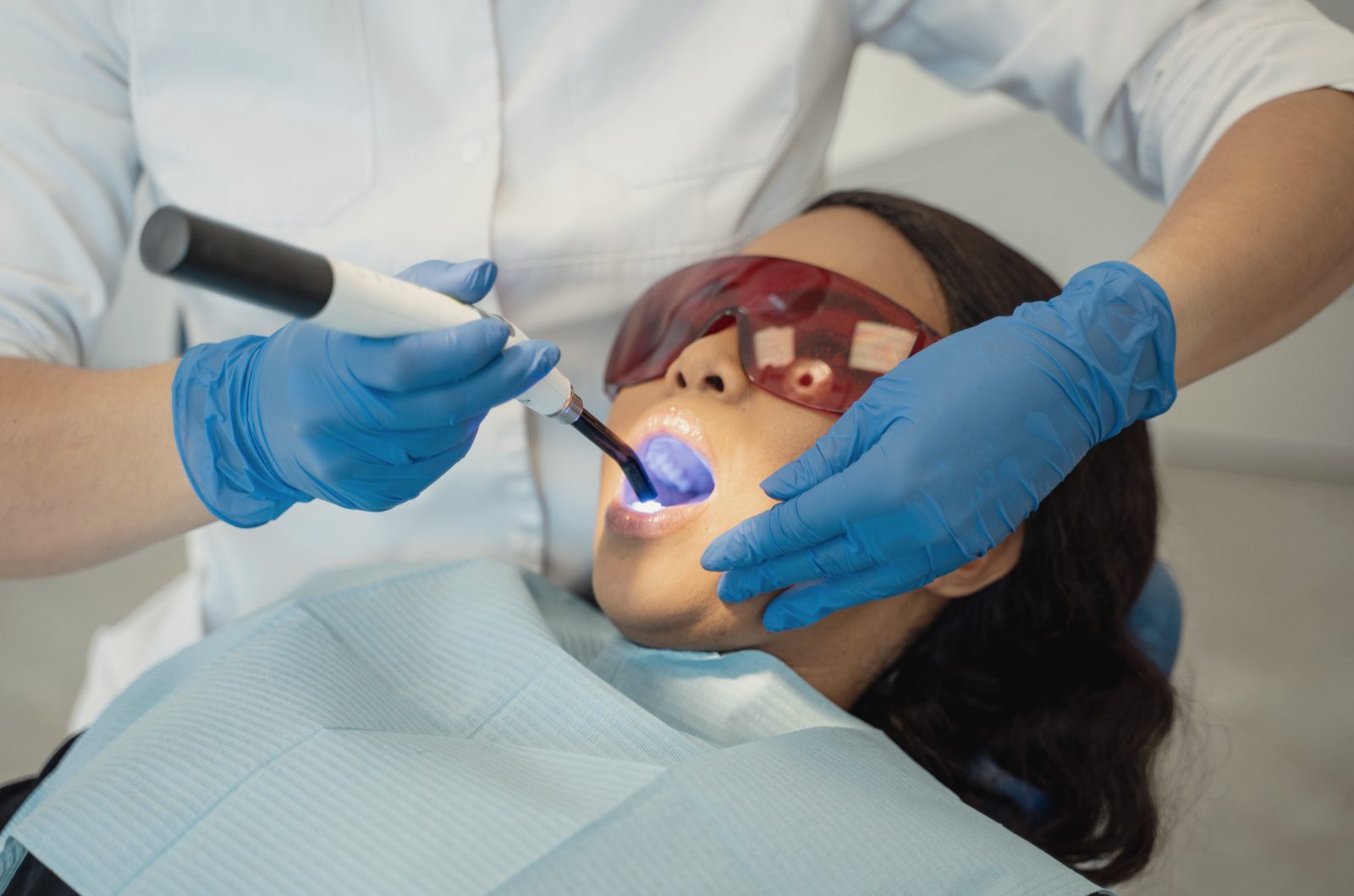 A dentist uses a small blue curing light on a patient’s tooth while the patient wears tinted safety glasses