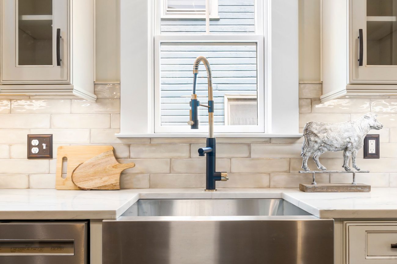 Finished kitchen: a stainless farmhouse sink under a window, white quartz counters, a tiled backsplash and glass-front upper cabinets