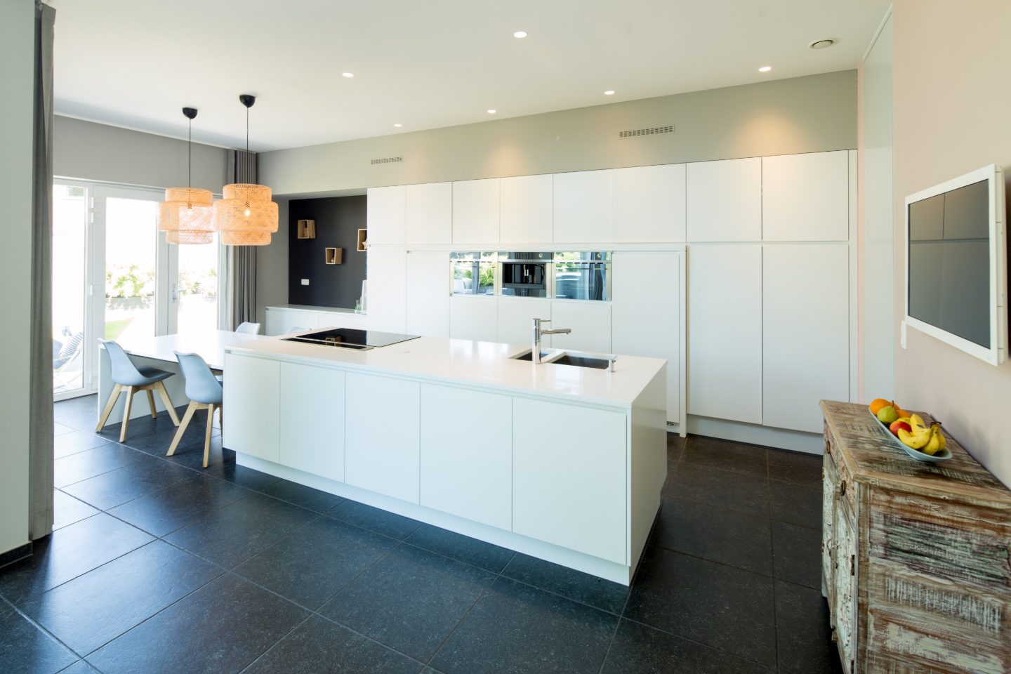 Remodeled kitchen with a long white quartz island, flat white cabinets from floor to ceiling, two woven pendant lights over a dining table and dark stone floor tiles