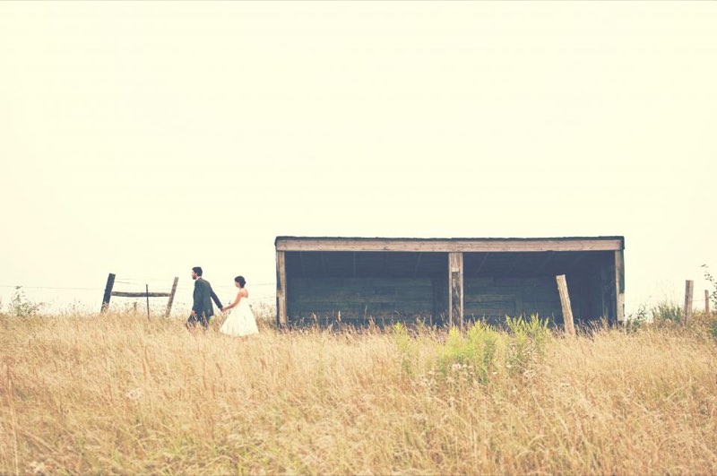 A couple walking hand in hand across a dry field toward an old barn
