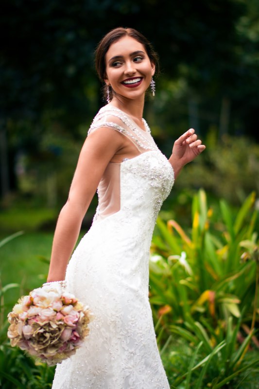 A bride smiling over her shoulder in a garden, holding a small bouquet