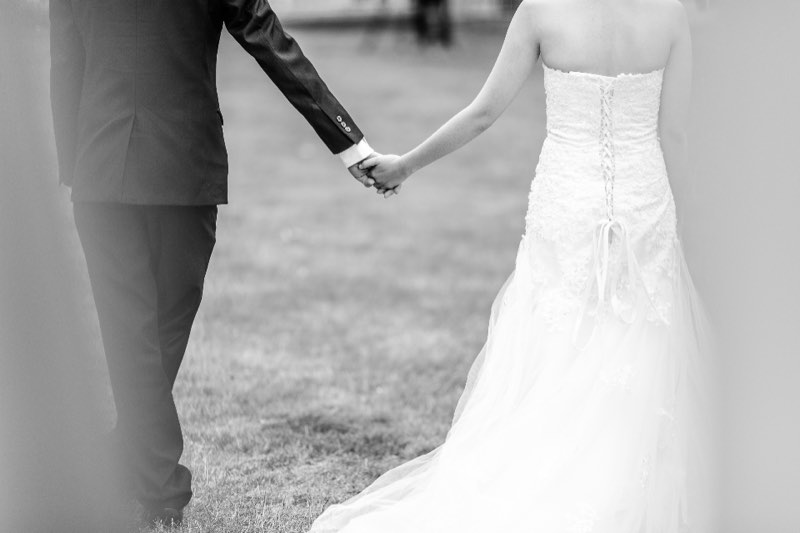 Black and white photo of a bride and groom walking hand in hand on grass