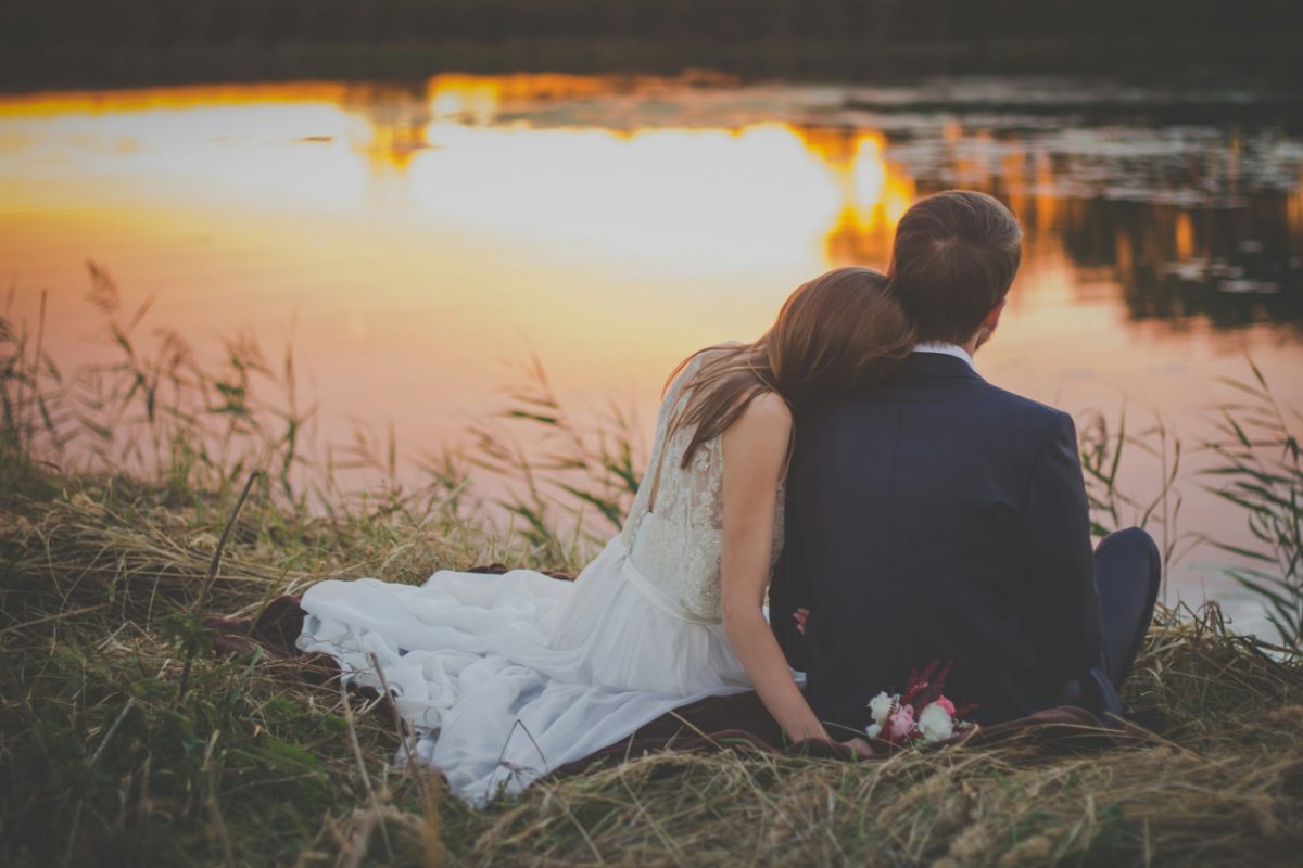 A couple sitting in tall grass at the water's edge, watching the sunset
