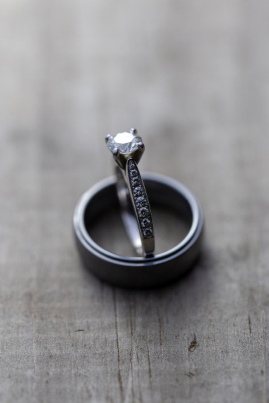 A round diamond solitaire with small diamonds set along its band, standing upright inside a dark men’s wedding band on weathered wood