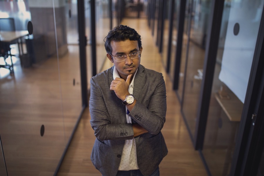 Ravi Vale in a gray jacket, standing in an office hallway with glass walls.