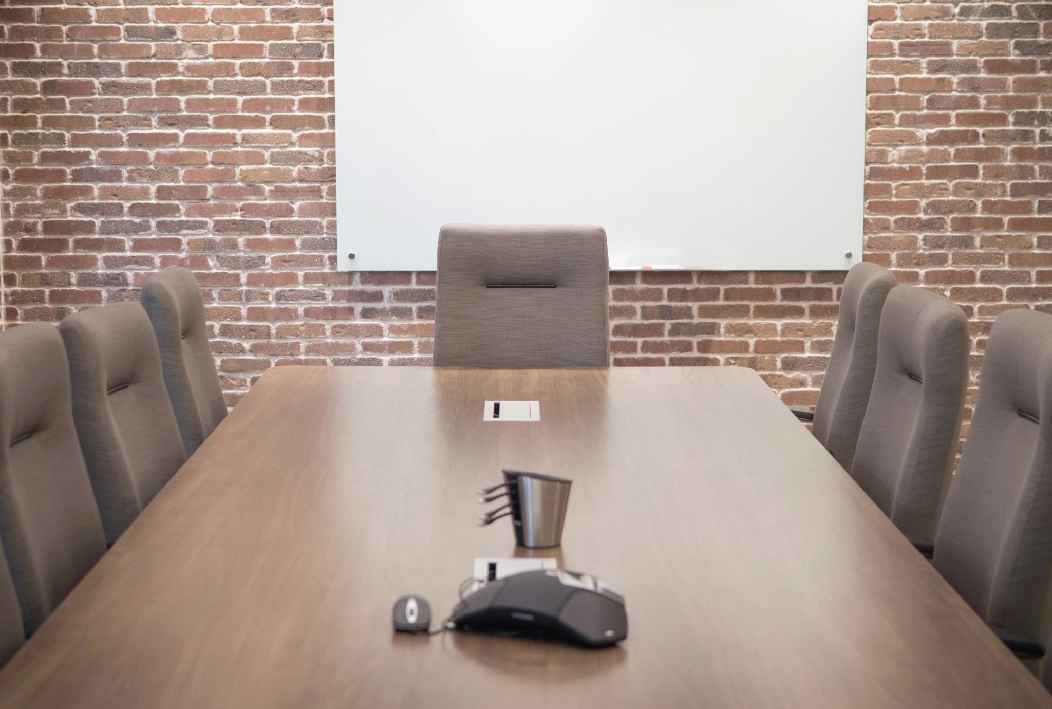 A conference room with a brick wall, a glass whiteboard, a long wooden table and grey upholstered chairs.