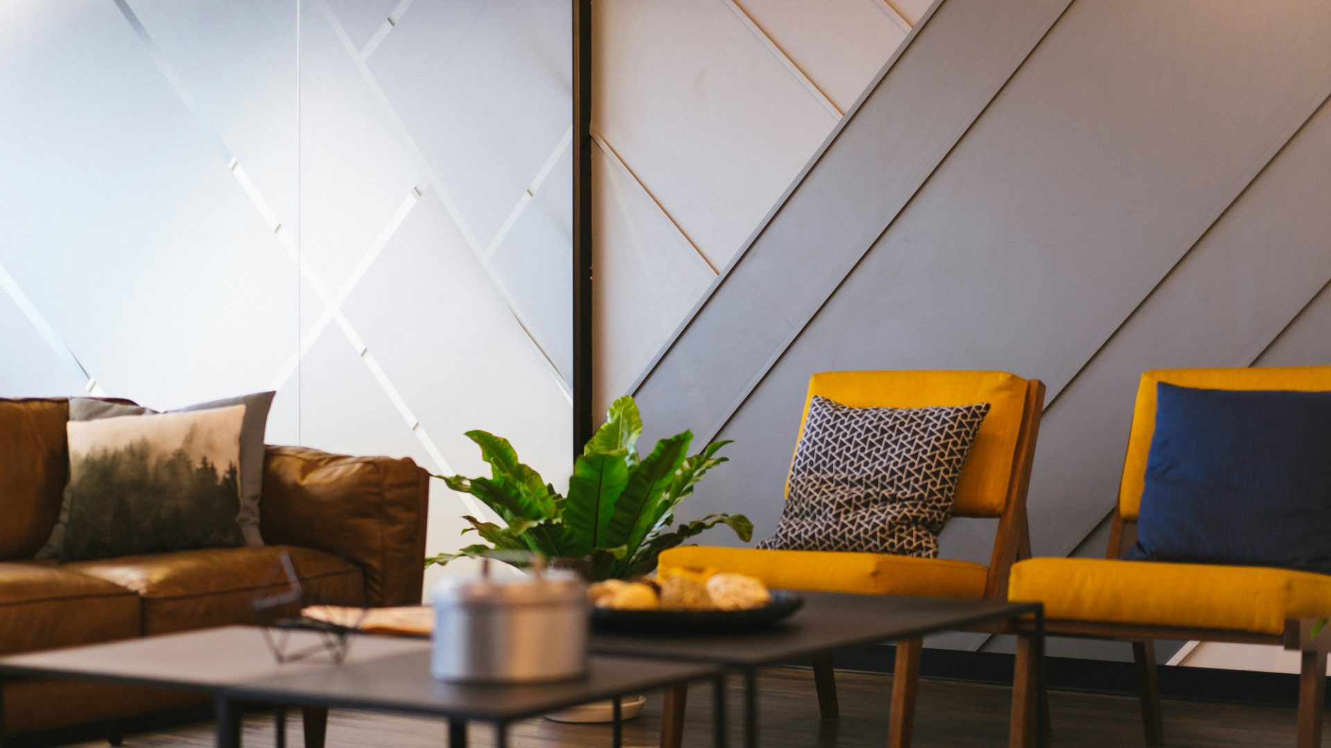 Family room with a cognac leather sofa, two mustard wool armchairs, a low black table with a large fern, against a wall of angled pale panels
