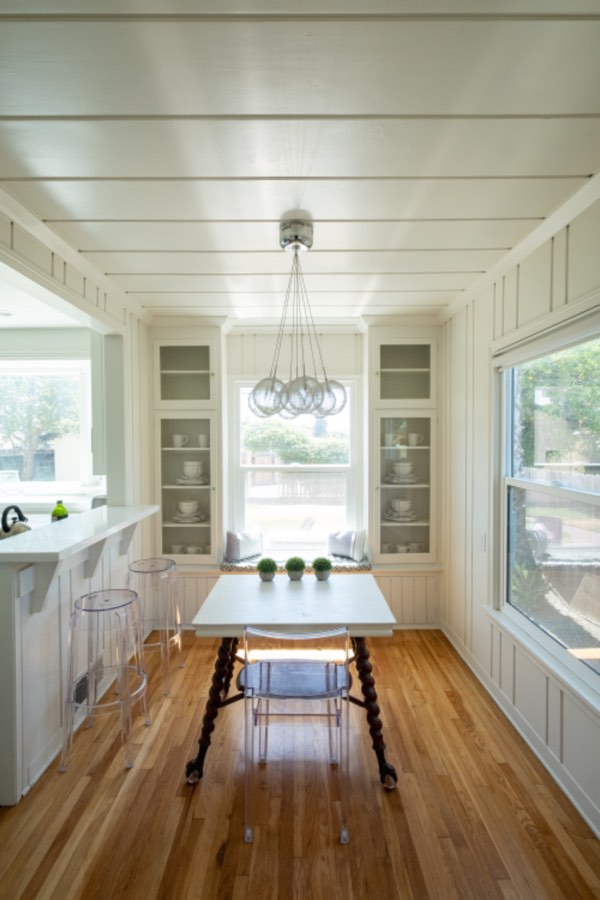 Bright breakfast nook with white board walls, glass-front cabinets on both sides of a window seat, a glass chandelier and a table with turned legs