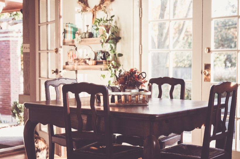 Sunlit dining room with a dark wood table, carved chairs with rush seats, and open shelves of plants and pottery between French doors