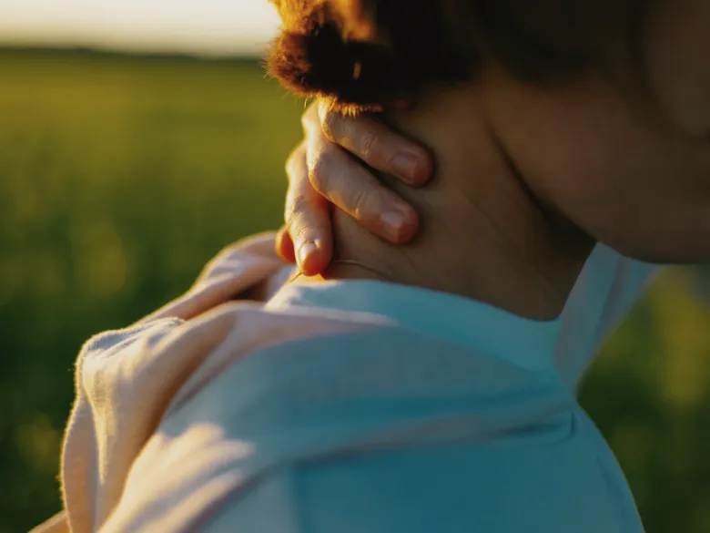 A woman in a pale blue shirt rubbing the back of her neck in a sunlit green field