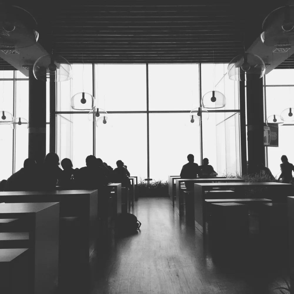 People in silhouette at long tables in a hall with a wall of windows