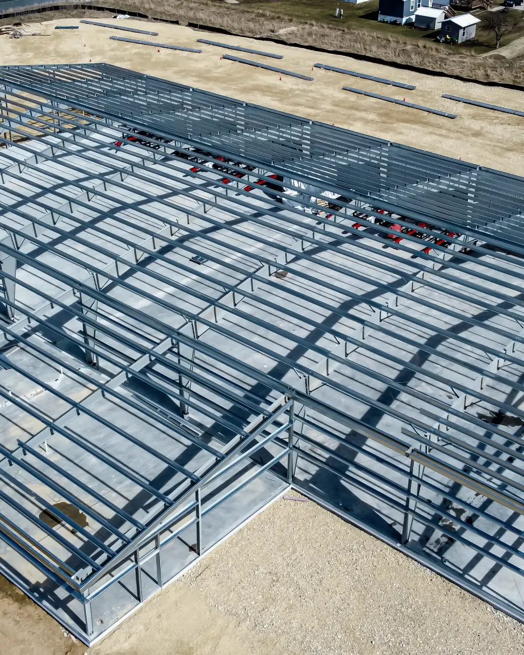 Aerial view of a long, low steel frame going up on a cleared site, its purlins casting a grid of shadows.