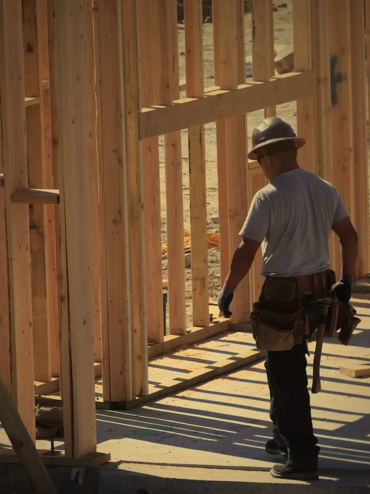 A crew lead in a hard hat and tool belt walks between newly framed walls.