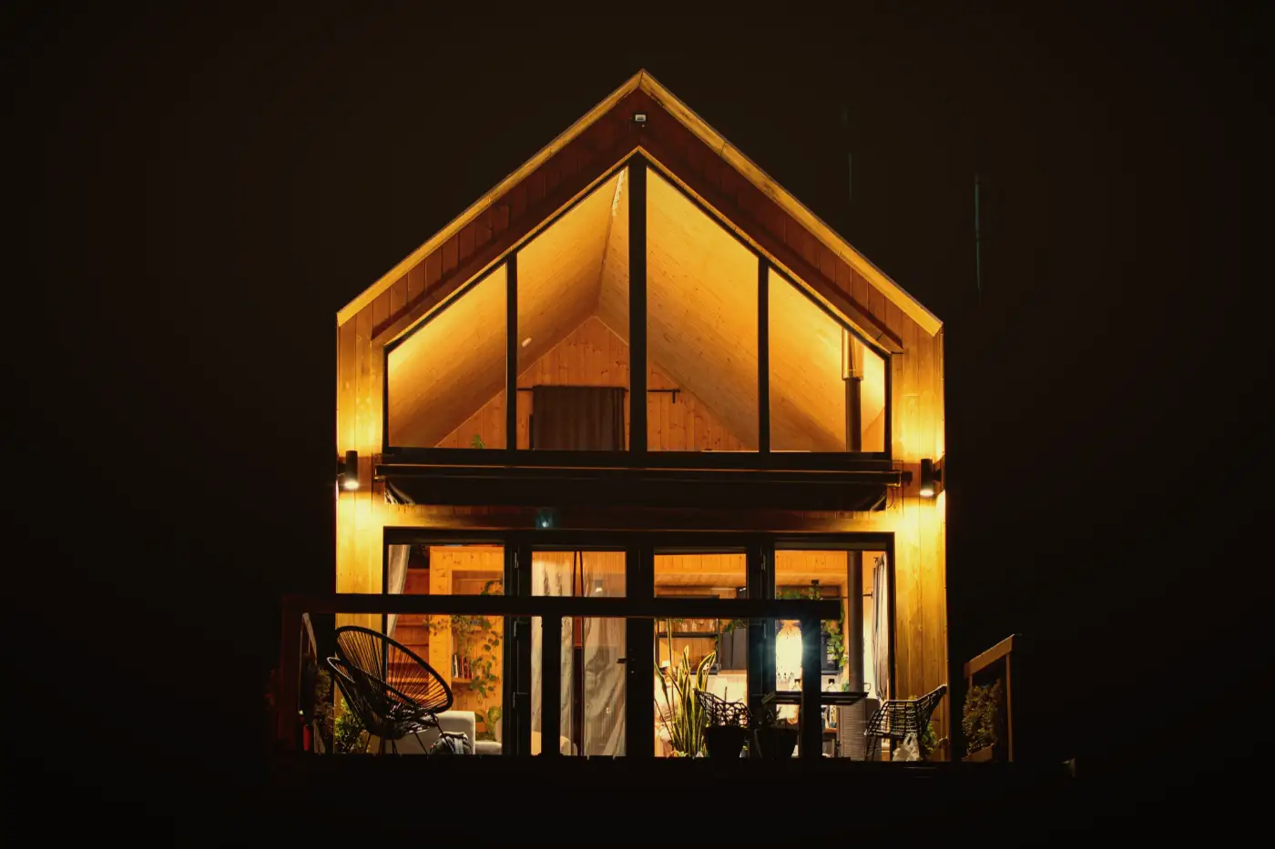 A modern timber-clad home with a tall glass gable, lit up at night above its deck.