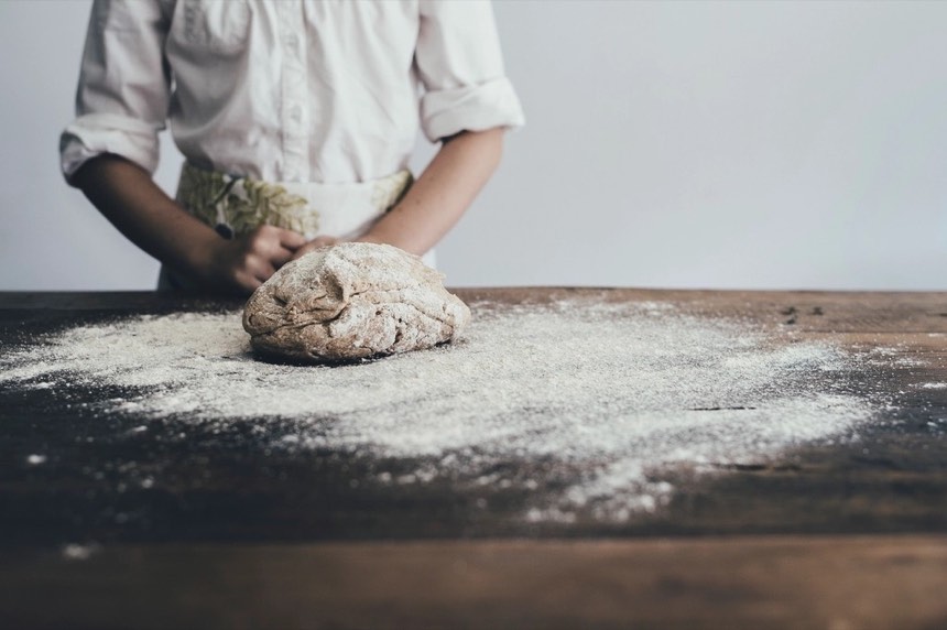 A baker in a white shirt and apron with her hands on a round of rye dough on a flour-dusted wooden table.