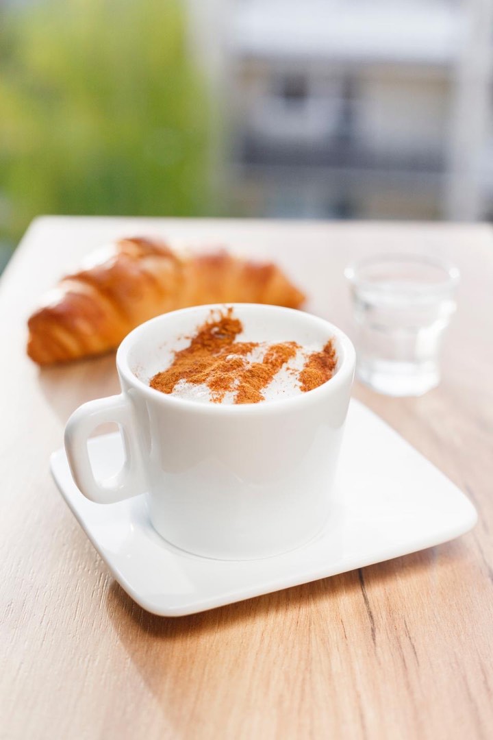 A cappuccino dusted with cinnamon on a white saucer, with a croissant and a glass of water behind it on a wooden table.