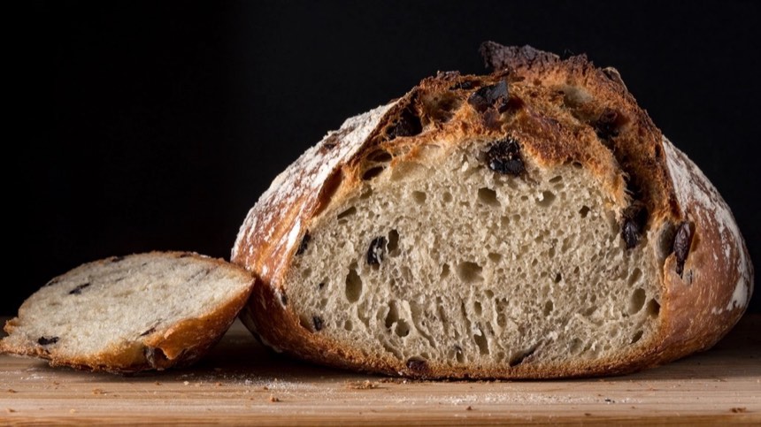 A crusty olive loaf cut open to show an airy crumb dotted with black olives, with one slice beside it.