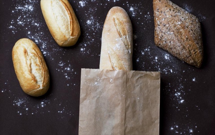 Crusty rolls, a seeded roll and a baguette in a brown paper bag on a dark, flour-dusted surface.