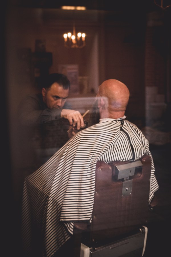 Gil cutting a client's hair, seen through the shop's front window in the evening
