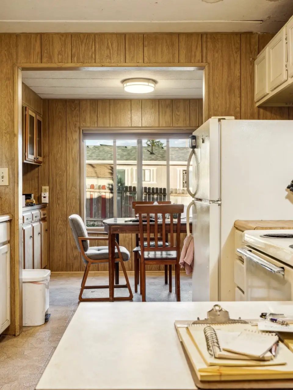 Sample before: a dated kitchen with wood-paneled walls, a white fridge, laminate counters and a small dining table by the window.