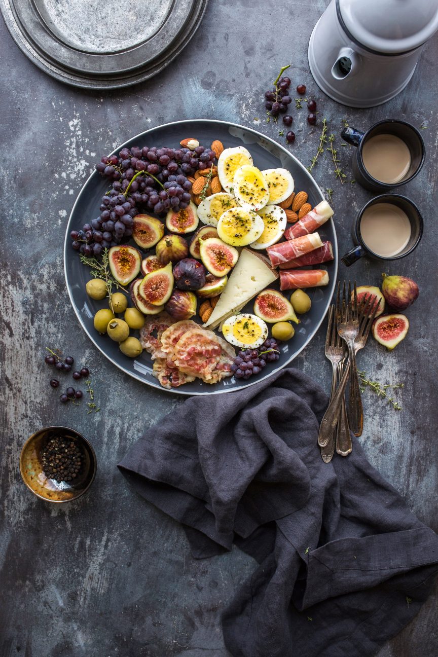 Overhead view of a slate table: a platter of halved figs, red grapes, soft-boiled eggs, prosciutto, cheese and green olives, with two cups of coffee beside it.
