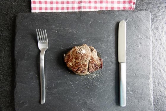 A seared steak on a slate board between a fork and knife, with a red gingham napkin at the top.