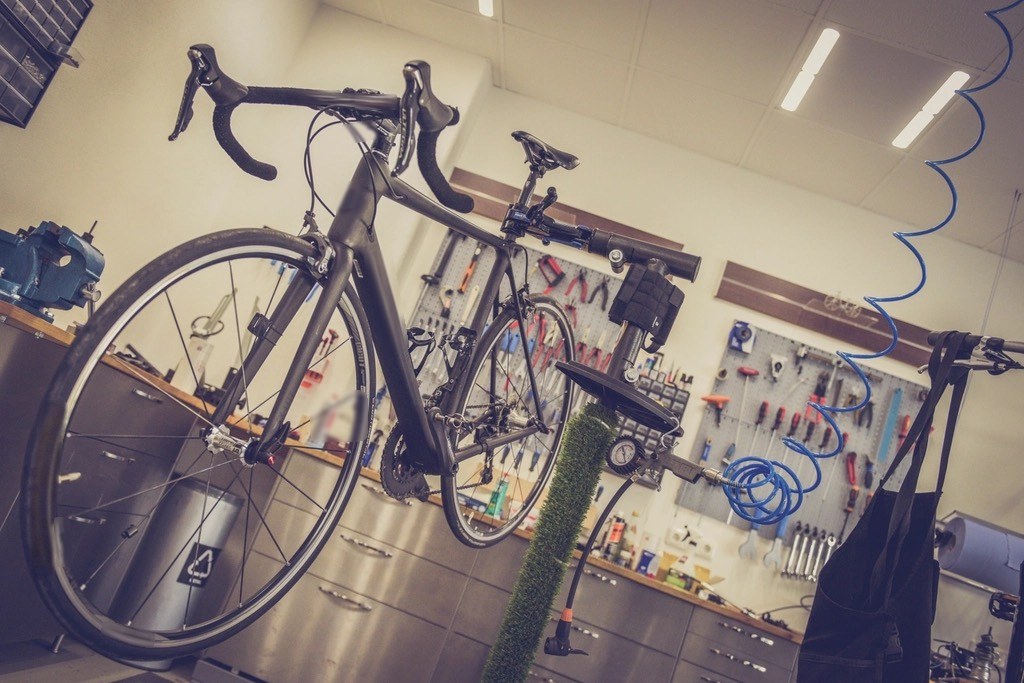 A black road bike clamped in a repair stand in a workshop, with tools hanging on pegboards behind it