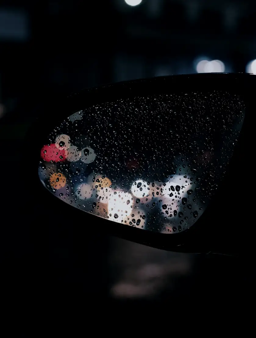 Rain beading on a coated side mirror at night, city lights behind