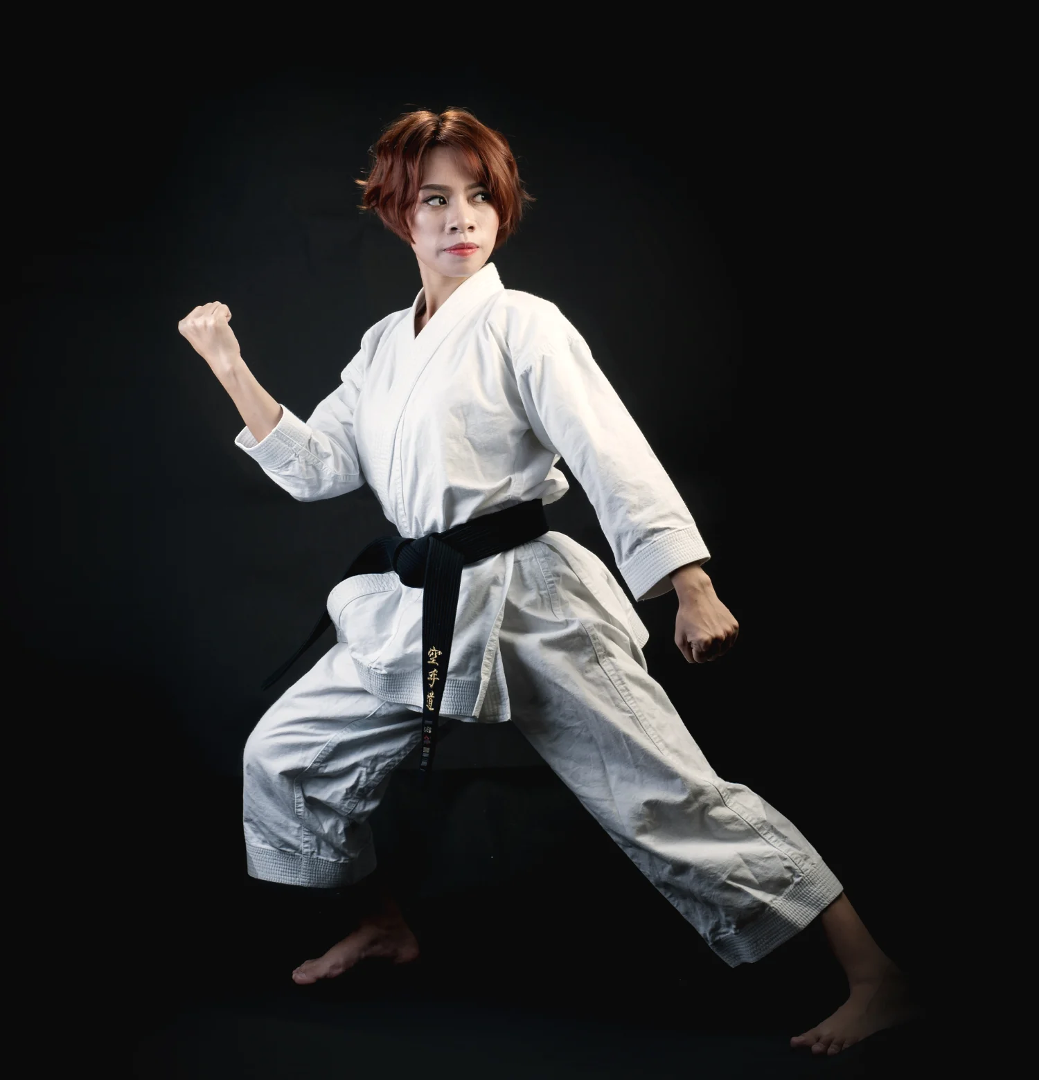 A karateka in a white gi and black belt holds a low stance, fist drawn back, against a black backdrop