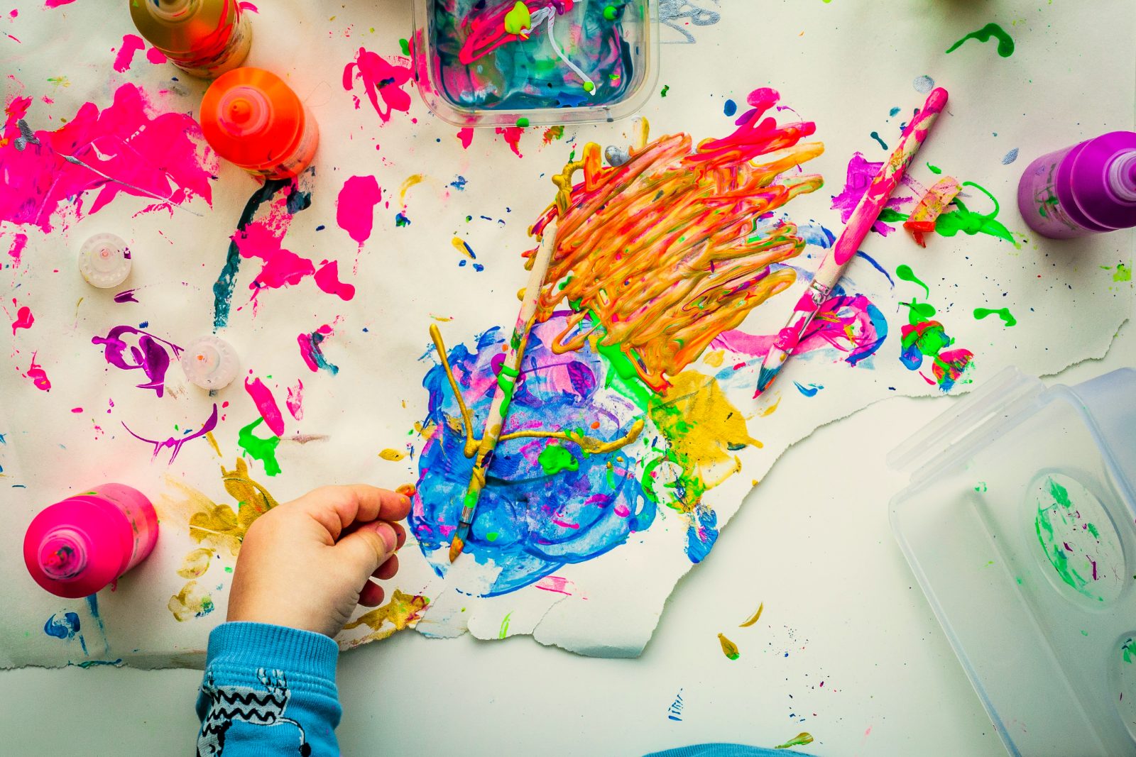 A child's hand reaches across paint-splattered paper on the art table, next to brushes and bottles of paint.