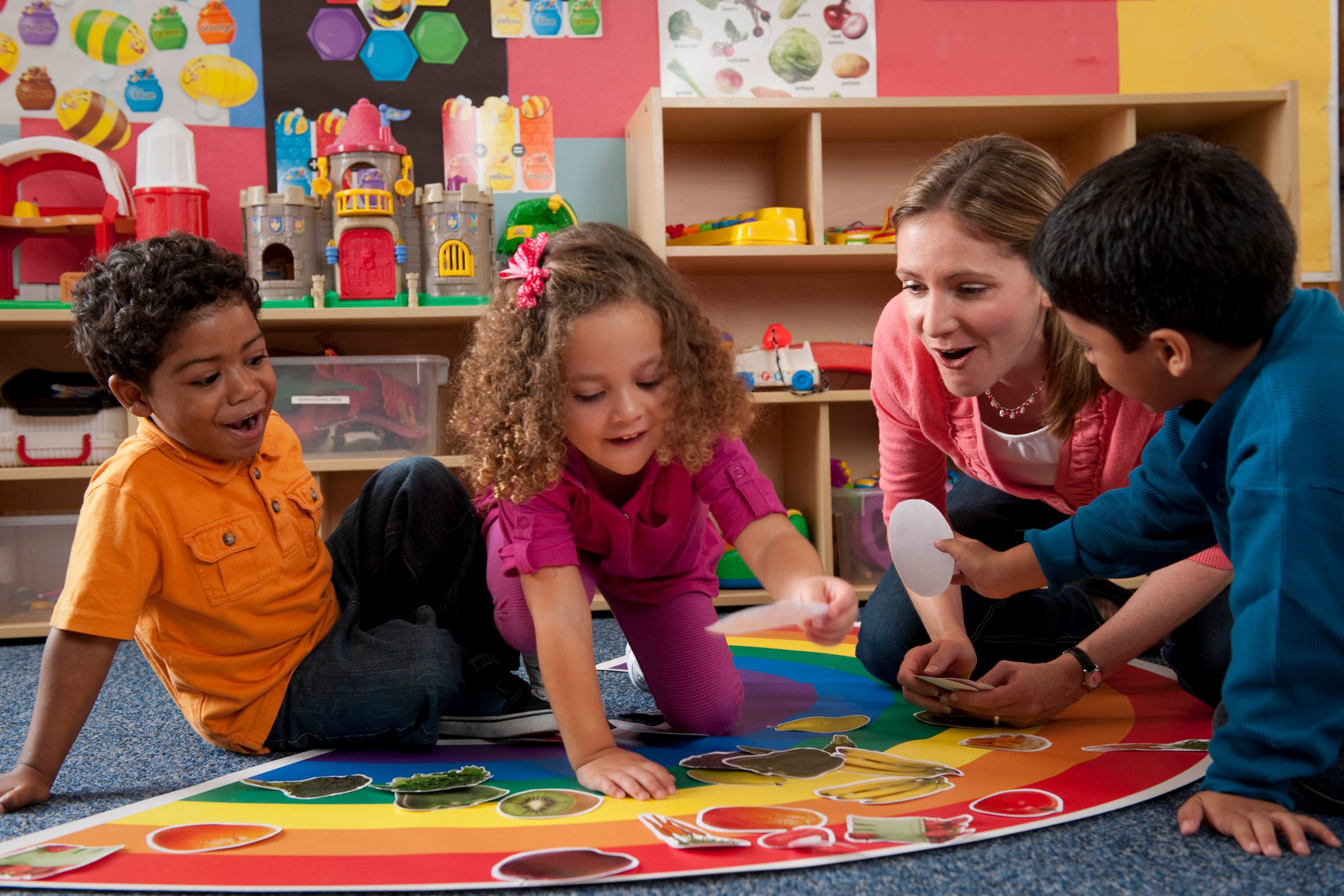 Four preschoolers and a teacher sit on a rainbow rug, sorting picture cards together in a bright classroom.