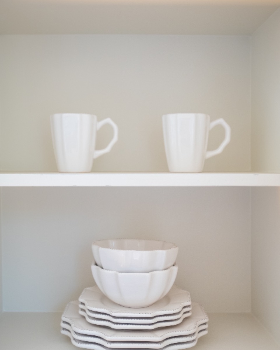 Open white cupboard with two mugs on the top shelf and stacked bowls and plates below