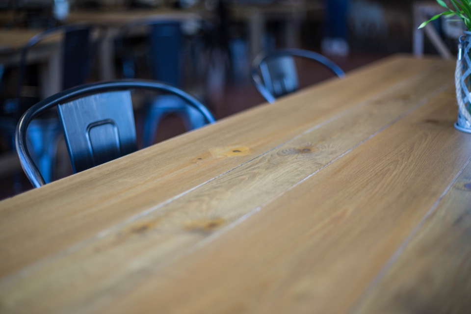 Close-up of a long, pale wood table top in a café, with metal chairs behind it