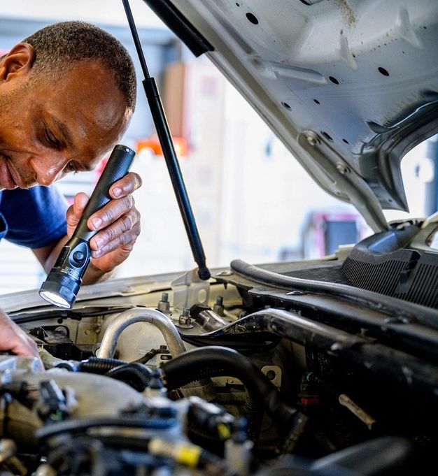 A smiling technician leaning into an engine bay with a flashlight