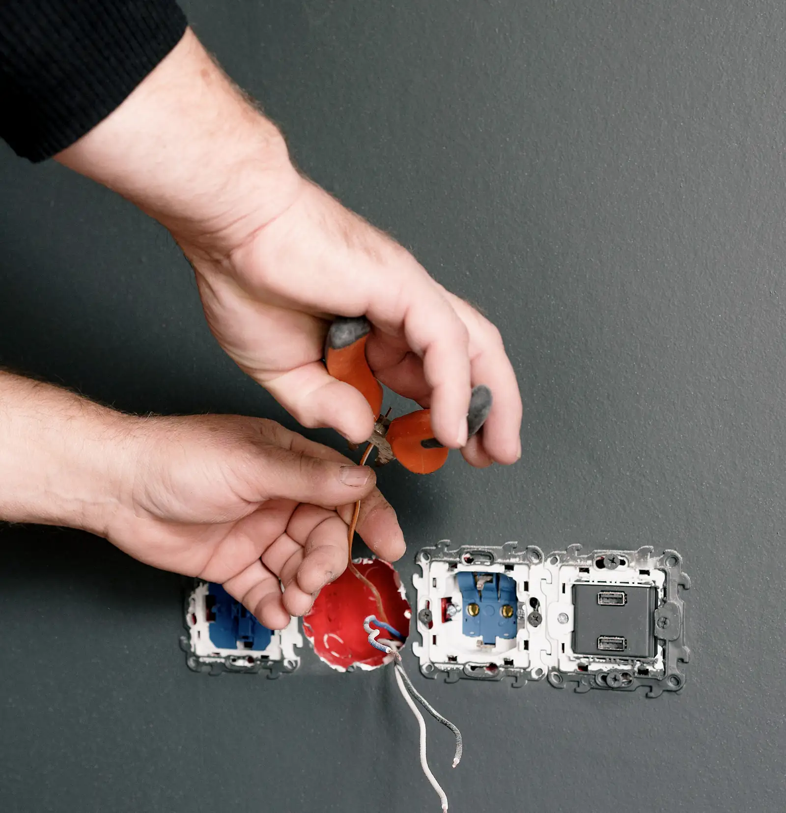 A Kestrel electrician trims a wire with pliers before landing it in a new device box on a charcoal wall.