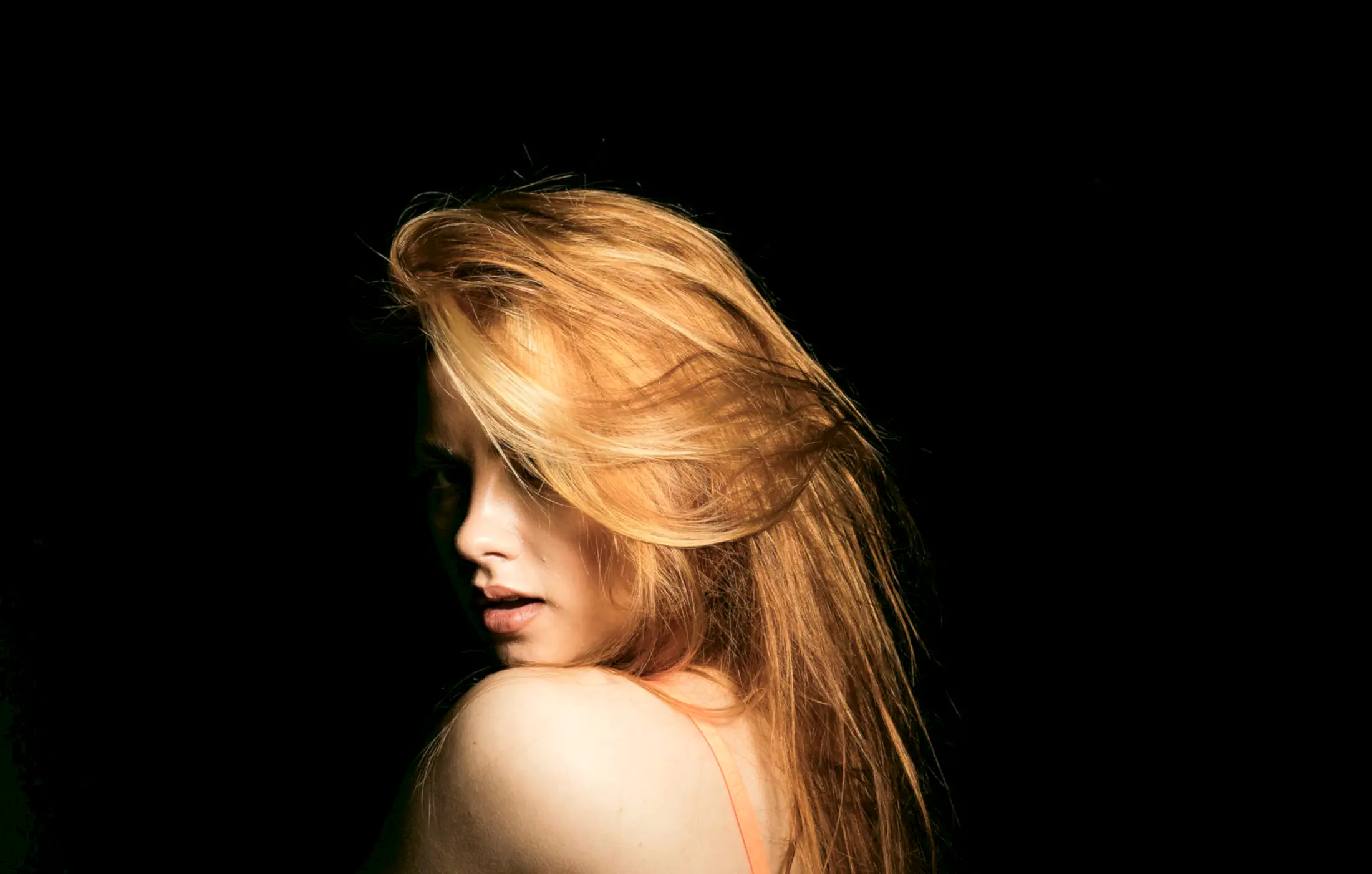 Long copper hair swept over a bare shoulder against a black backdrop.