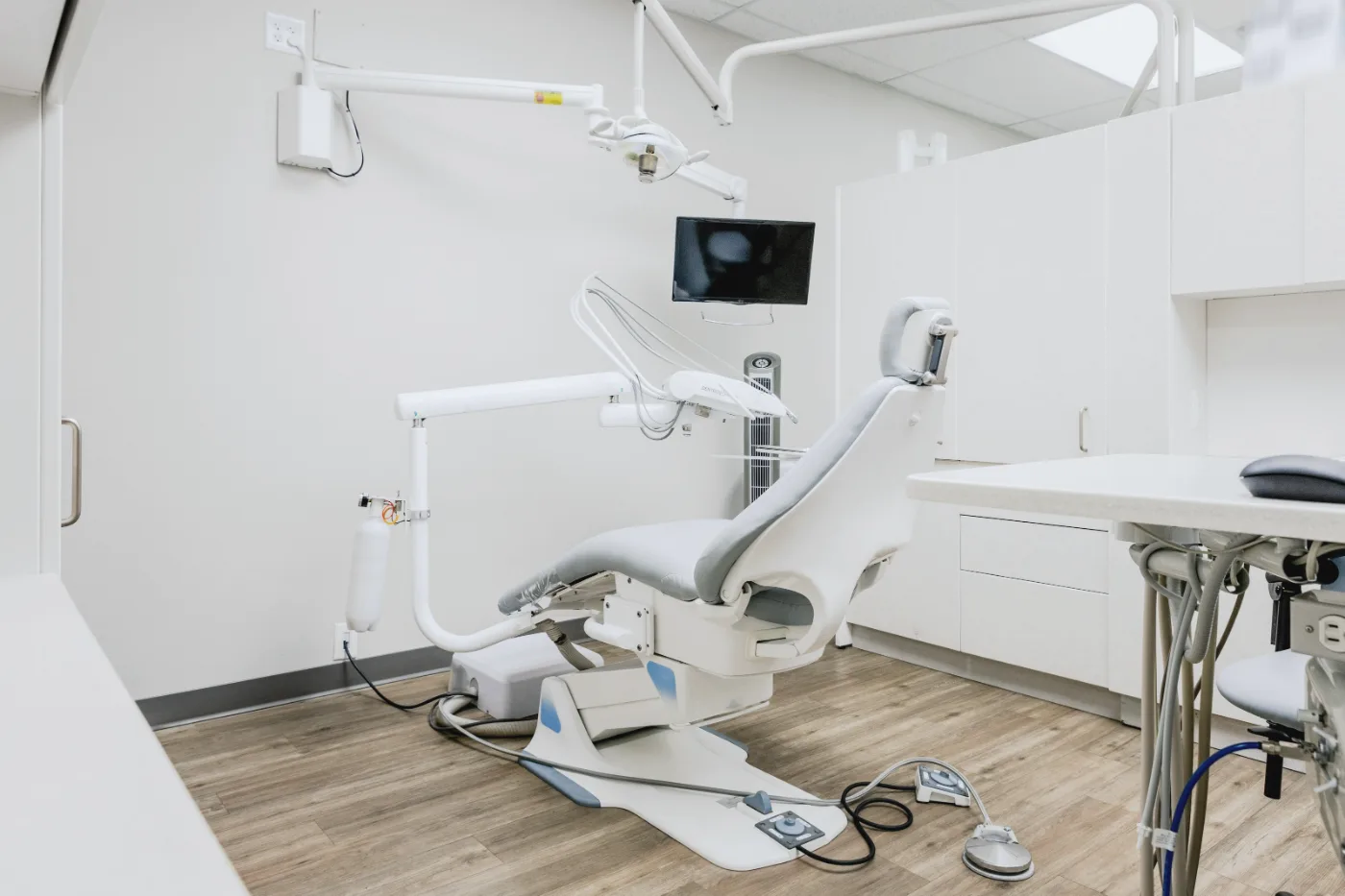 A bright treatment room with a pale grey dental chair, white cabinets and a wood floor