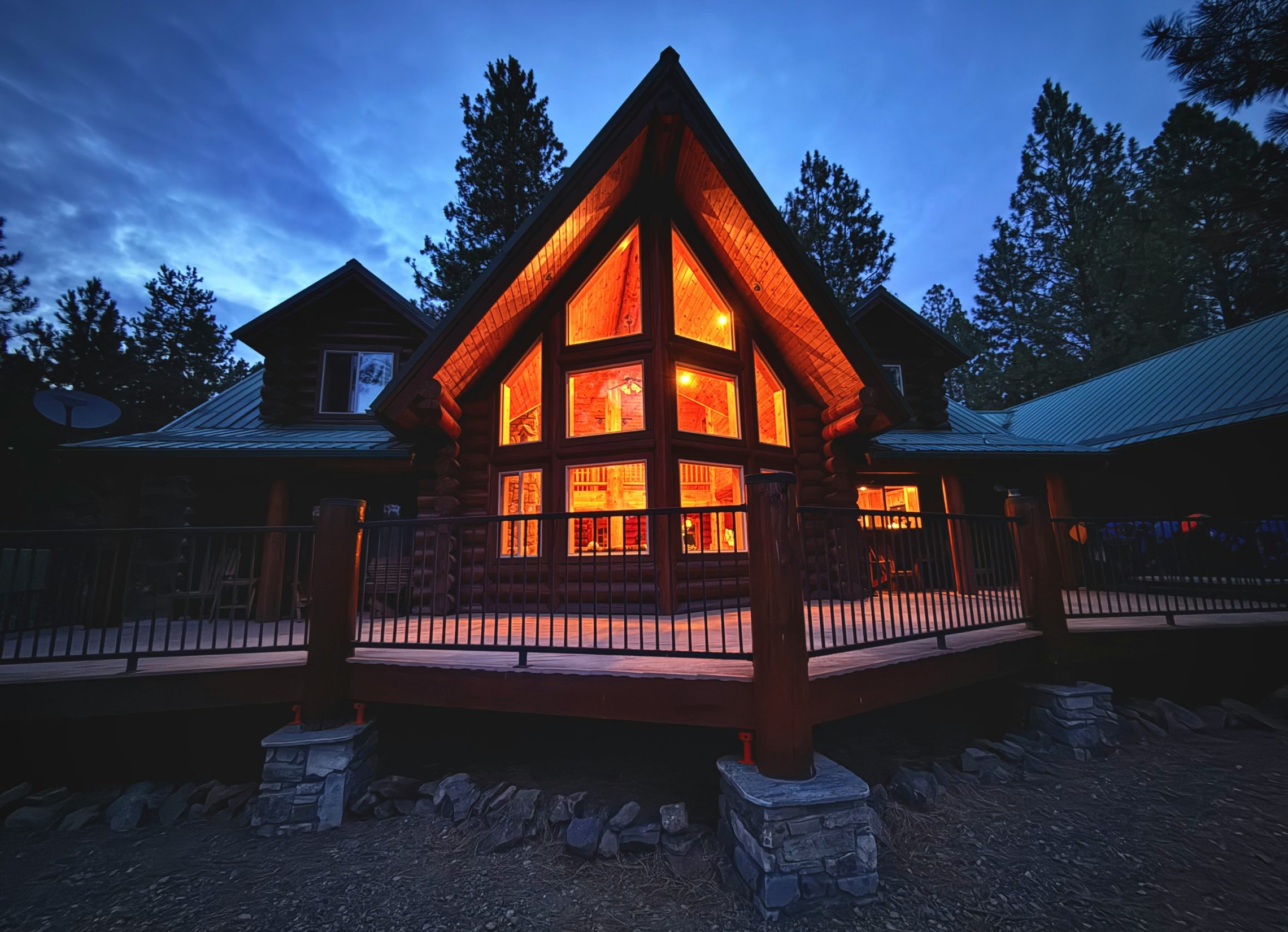 Log house at dusk with a tall glass gable and a wraparound deck, lit warmly from inside