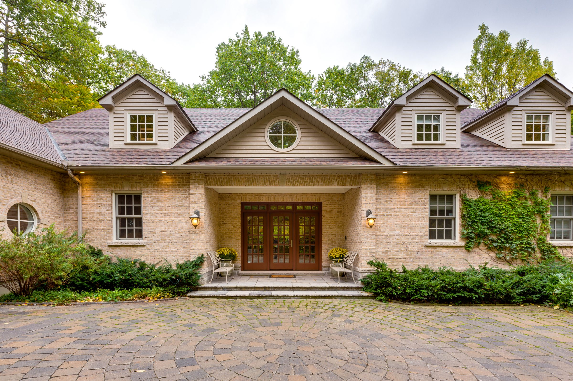 Stone house with three dormers, a round window and double wooden doors above a curved paver driveway