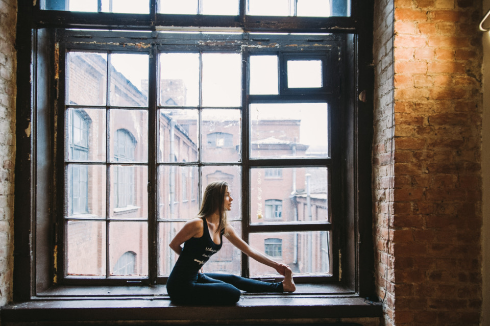 A student stretching on the floor beside a tall arched window in the studio's brick-walled room